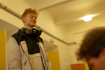 white student smiling in dorm corridor, headphones around neck, warm fluorescent lighting, relaxed confident pose, casual jacket and youthful energy suggesting peer mentorship