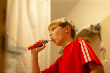 White boy brushing teeth in bathroom mirror, red shirt, electric toothbrush foam, focused morning routine, preparing for school with confident expression, warm bathroom lighting