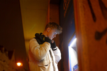 Young white man at atm kiosk withdrawing cash under neon light, wearing white puffer jacket and headphones, gloved hands entering card, urban street background, cold evening atmosphere,