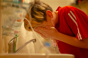 young person unknown ethnicity splashing face at sink after practice, red sports shirt damp with sweat, towel on shoulder, fluorescent lockerroom light, leaning over basin, water droplets