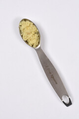 One teaspoon of light yellow, freshly grated ginger is shown inside a metal measuring spoon placed diagonally on a plain white surface