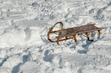 A classic wooden sled with a woven seat sits on a field of deep uneven snow in the bright winter sunlight