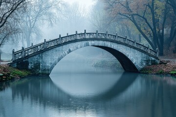 Fototapeta premium Serene stone bridge over still water in a foggy park landscape during autumn morning