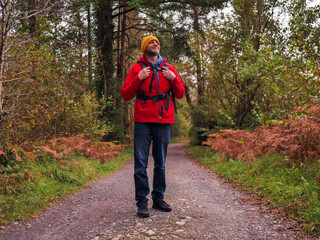 Obraz premium A man wearing a red jacket and a yellow hat and black backpack is standing on a dirt road in a forest.. Travel and outdoor adventure theme. Male with grey beard in his 50s.