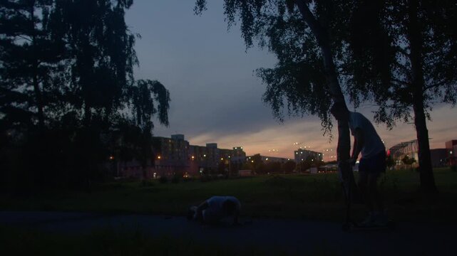 male athlete city park sunset collapsed on path while partner kneels to assist, twilight skyline and tree silhouettes frame urgent but calm outdoor aid scene, focus on support and recovery