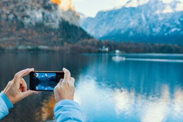 A female traveler holds a smartphone to capture the stunning reflection of the Hallstatt mountains and lake during a crisp autumn day