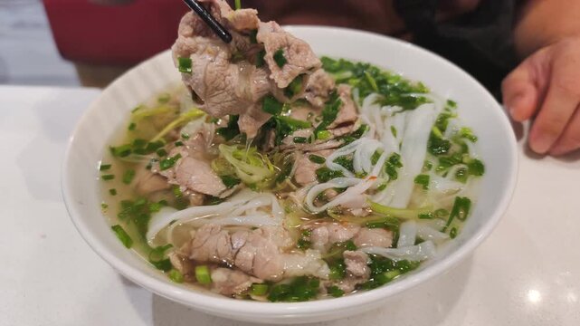 Delicious vietnamese pho bo for lunch on white table. Person eats vietnamese pho bo with chopsticks. This vietnamese pho bo is authentic cuisine with rice noodles and sliced meat and greens.