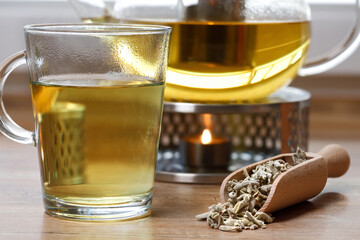 Sage tea in a glass next to dried leaves in a wooden spice spoon on the table with a glass teapot with warmer and tea light, concept for naturopathy and healthy homeopathic herbs