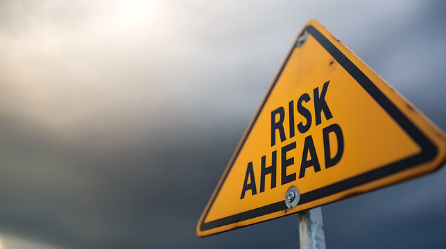 A yellow, triangular sign warns of 'Risk Ahead' against a backdrop of a cloudy sky. The sign is mounted on a pole, alerting passersby to potential danger or uncertainty ahead.