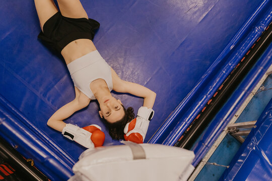 Top view of a tired young woman boxer in red gloves lying on the blue floor of a boxing ring and looking at camera after intense workout, exhaustion and female determination concept