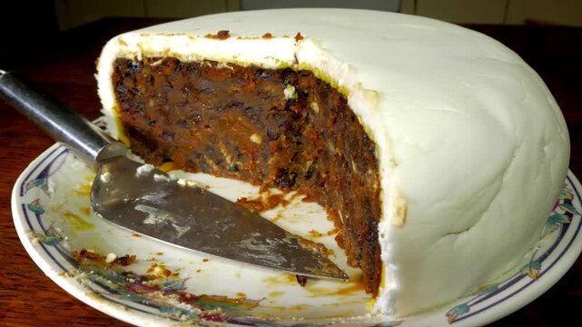 Closeup circular dolly shot of a traditional, partly eaten, fruit filled, Christmas cake, topped with a layer of marzipan and icing sugar, resting on a plate with a slicing tool, on a kitchen table.
