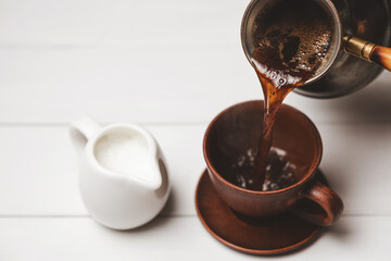 Pouring Turkish coffee from cezve into ceramic cup with milk pitcher on white rustic table