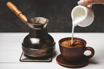 Female hand pouring milk into coffee cup next to Turkish cezve on white rustic table