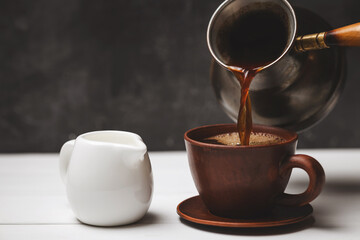 Pouring Turkish coffee from cezve into ceramic cup with milk pitcher on white rustic table