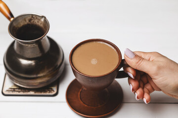 Close-up of female hands holding ceramic coffee cup next to Turkish cezve on white rustic table