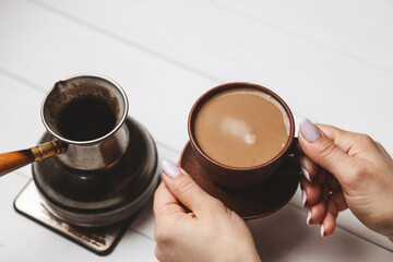 Close-up of female hands holding ceramic coffee cup next to Turkish cezve on white rustic table