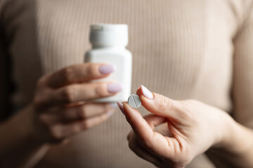 Close-up of female hands holding white pill bottle and single white round pill with score line,...
