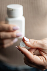 Close-up of female hands holding white pill bottle and single white round pill with score line, concept of medication and pharmaceutical treatment