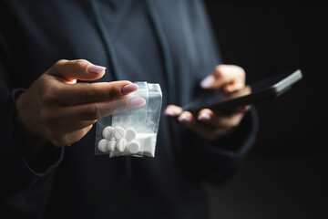 Close-up of female hands holding smartphone and two transparent plastic bags with white narcotic powder and white pills, concept of drug abuse and drug trafficking