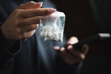 Close-up of female hands holding smartphone and two transparent plastic bags with white narcotic powder and white pills, concept of drug abuse and drug trafficking