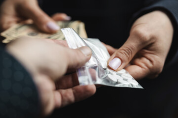 Close-up of addicted person hands paying cash to buy two transparent bags with narcotics, white pills and powder from drug dealer, concept of drug abuse and drug trafficking