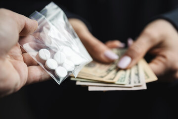 Close-up of addicted person hands paying cash to buy two transparent bags with narcotics, white pills and powder from drug dealer, concept of drug abuse and drug trafficking