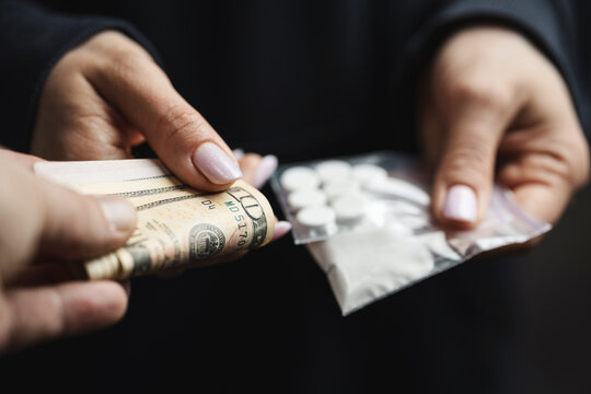 Close-up of addicted person hands paying cash to buy two transparent bags with narcotics, white pills and powder from drug dealer, concept of drug abuse and drug trafficking