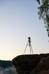A photographer&rsquo;s tripod and camera stand silhouetted on a rocky outcrop, ready to capture the tranquil moment. Atmospheric sunrise over a misty lake surrounded by forested hills.