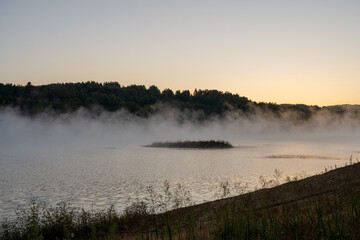 Morning fog looms over Lake Vlasina, revealing a small island with lush vegetation against a backdrop of dark forest. Warm sunlight illuminates the fog and the surface of the water.