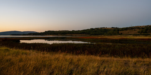 Minimalist dusk landscape featuring tall dry meadow grass, a reflective lake, and distant forested...