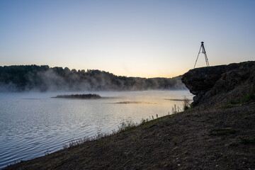 A photographer&rsquo;s tripod and camera stand silhouetted on a rocky outcrop, ready to capture the tranquil moment. Atmospheric sunrise over a misty lake surrounded by forested hills.