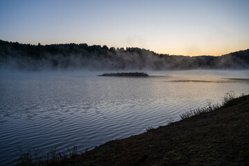 Golden morning mist rises from the calm waters of Lake Vlasina in Serbia, surrounding a small vegetated islet with a forested shoreline in the background.