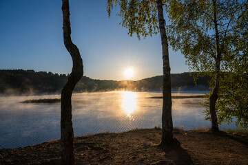 Morning fog looms over Lake Vlasina, revealing a small island with lush vegetation against a backdrop of dark forest. Warm sunlight illuminates the fog and the surface of the water.