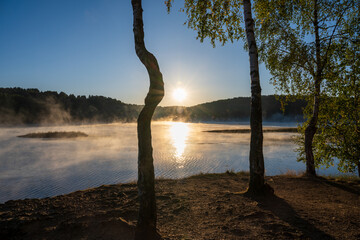 Golden morning mist rises from the calm waters of Lake Vlasina in Serbia, surrounding a small vegetated islet with a forested shoreline in the background.