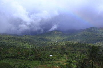 Naklejka premium morning in the mountains. The image is a scenic photograph of lush, green mountains in a tropical location, likely within the Philippines, with a rainbow visible in the upper right. 