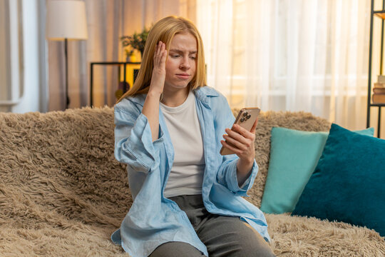 Young woman sits at home holding smartphone, discovering bad news and feeling shock, sadness and regret. Caucasian girl collapses on couch, revealing despair after phone defeat fail under sunset glow