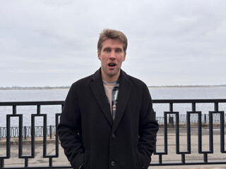 A man with light hair stands by the river, looking surprised while wearing a coat