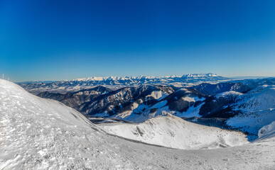 Wide panoramic view of snow-covered peaks in the Low Tatras National Park, Slovakia, featuring a bright sun star, frozen mountain ridges, and a distant view of the High Tatras under a clear winter sky