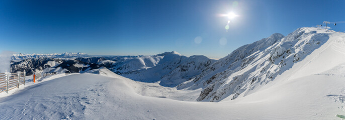 Wide panoramic view of snow-covered peaks in the Low Tatras National Park, Slovakia, featuring a bright sun star, frozen mountain ridges, and a distant view of the High Tatras under a clear winter sky