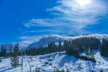 Wide view of a snowy ski resort area with evergreen pine forests and high mountain peaks in the background under a bright winter sun and blue sky in Slovakia.