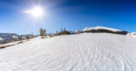 Wide panoramic view of a groomed ski slope in Jasna, Low Tatras, Slovakia, featuring a bright sun star in a clear blue sky and snow-covered mountain ridges in the background.
