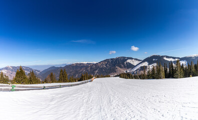 A wide panoramic view of a snow-covered ski slope in the Low Tatras National Park, Slovakia, featuring a wooden safety fence, evergreen trees, and distant mountain peaks under a clear blue sky.
