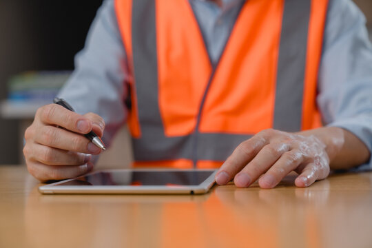 Engineer wearing safety vest using a tablet with stylus pen at a desk. Concept of digital inspection, construction technology, industrial workflow, and modern engineering work.