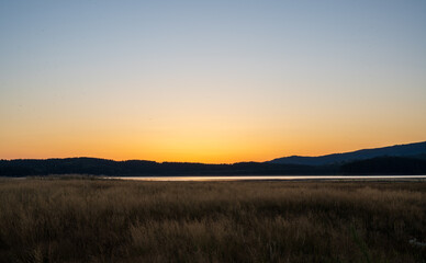 Golden sunset over a calm lake with wide open grassland in the foreground and dark rolling hills on the horizon. The clear sky fades from warm orange to soft blue.