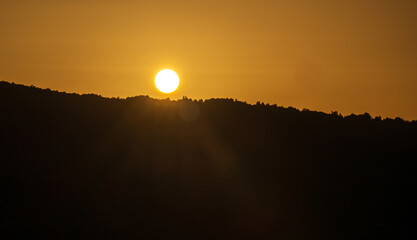 A golden sunset over calm lake waters with a reflective trail of light leading to the silhouettes of hills. Warm orange tones fill the sky, creating a serene twilight landscape ideal for sunset themes