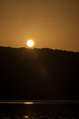 Golden hour over a quiet lake with the sun shining on reflective water and silhouetted hills on the horizon. Evocative sunset landscape with warm light and calm mood, suited for backgrounds.