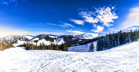 A scenic wide-angle panorama of a groomed ski slope in the Low Tatras, Slovakia, featuring ski lift infrastructure, evergreen forests, and dramatic cloudy sky over snow-covered mountain peaks.