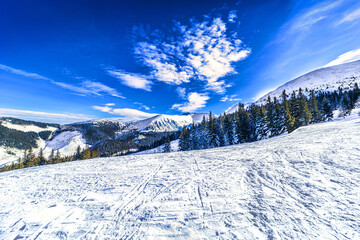 A wide scenic shot of a groomed ski slope in the Low Tatras, Slovakia, under a dramatic cloudy sky during twilight, featuring snow-covered ridges and evergreen forests.