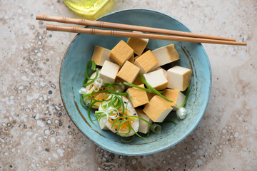 Smoked tofu with soy-marinated egg and spring onion in a turquoise bowl, horizontal shot on a beige granite background, high angle view