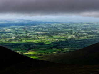 From a high vantage point on a mountain, one can see farmland, green fields, and small towns in Ireland. Dark clouds loom over the landscape.